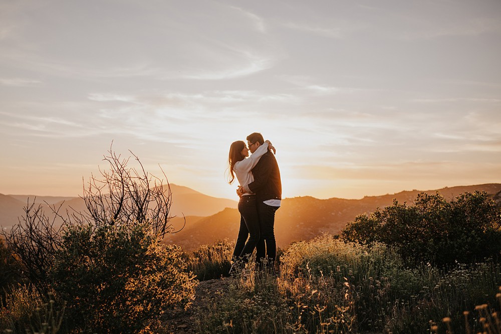 Couple embraces on a hilltop at sunset with mountainous scenery.