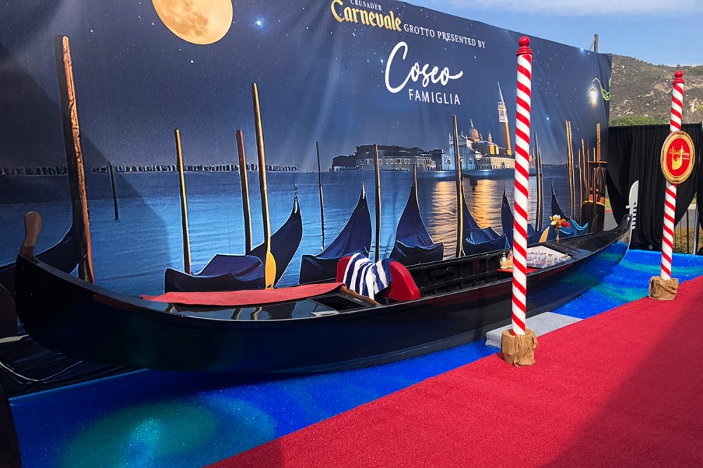 Black Venetian gondola displayed with a moonlit Venice backdrop and red carpet setup for an Italian-themed event or photo booth rental.