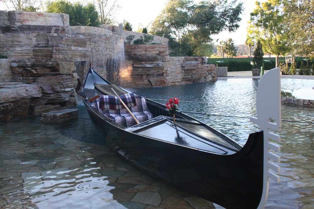 Elegant black Venetian gondola with blankets and flowers set up beside a stone waterfall in a pool, perfect for event decoration or photo prop rental.