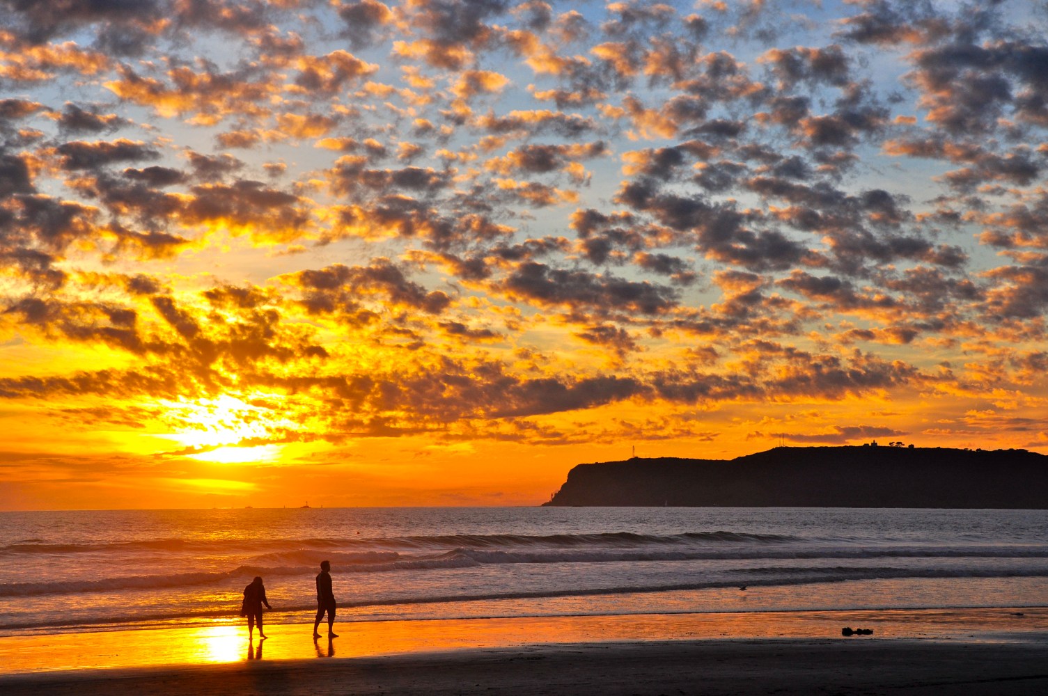 Two people walking on beach in Coronado at sunset with Point Loma in view.