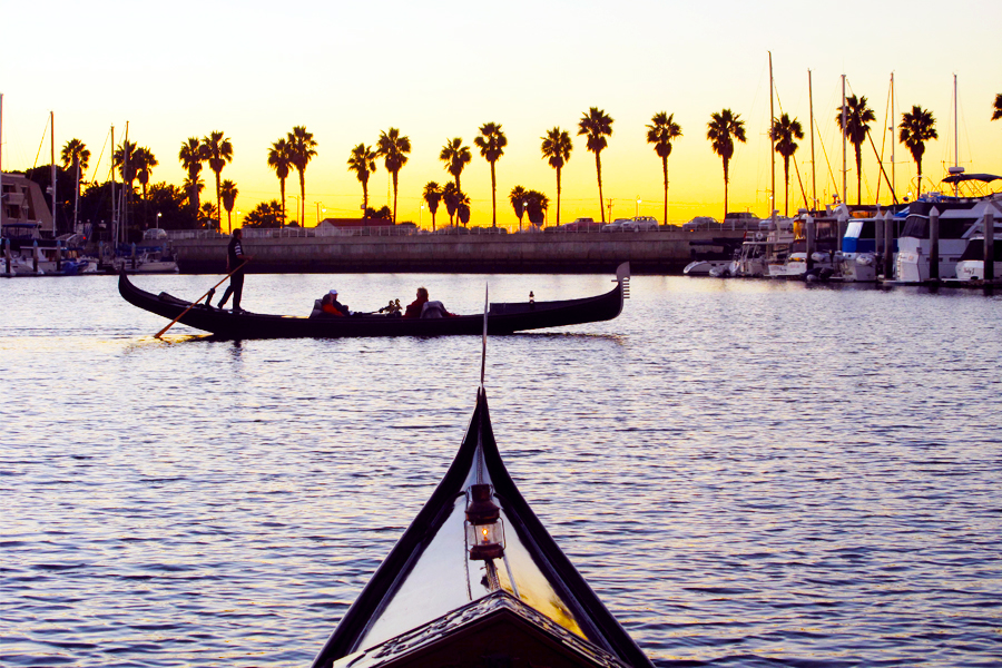 Gondola on water at sunset with palm trees and boats in background.