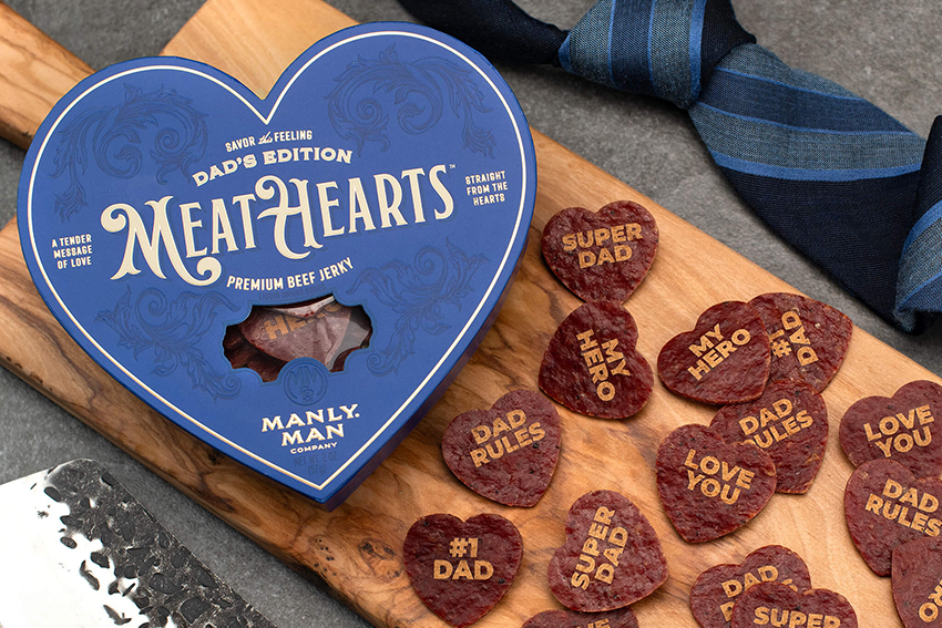 Heart-shaped beef jerky with messages, tie, and blue box on a board.